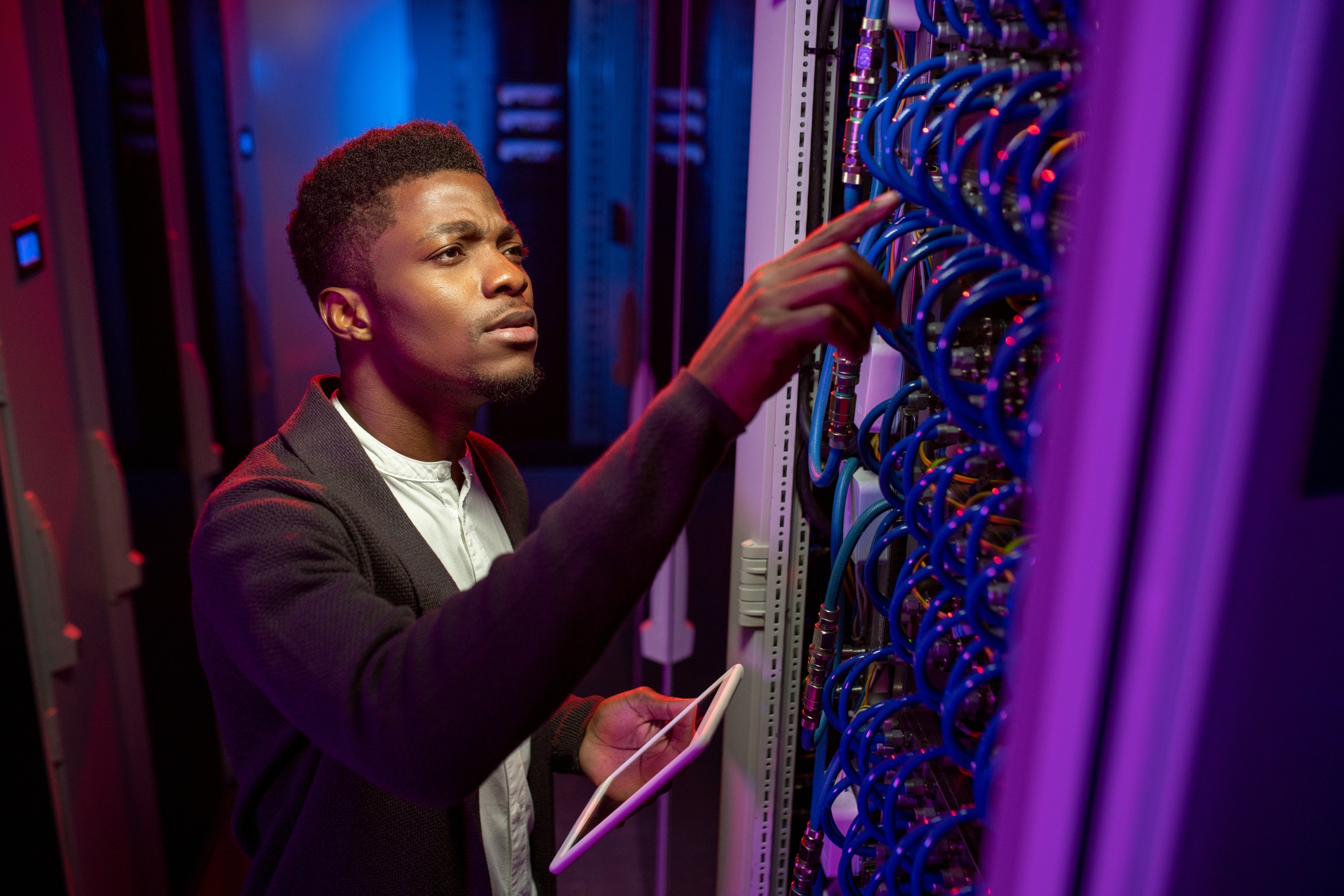 Concentrated IT engineer standing with tablet in data center and checking connection of cables for customer